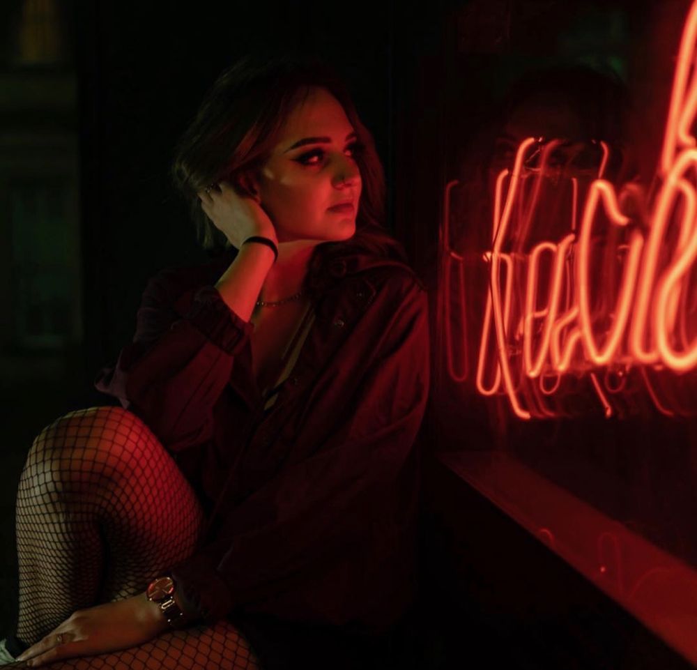 Maisie in a dark jacket and fishnets, lit red by a neon sign