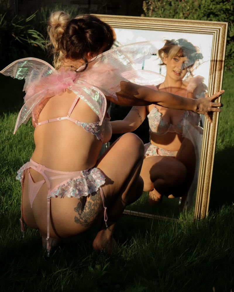 Maisie in pastel lingerie and iridescent fairy wings, crouched at a gold mirror in a garden